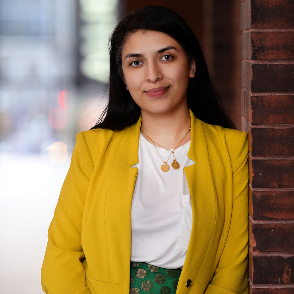 Photograph of Jaspreet Sandhu, a South Asian woman with long black hair. She is wearing a white blouse and yellow blazer.