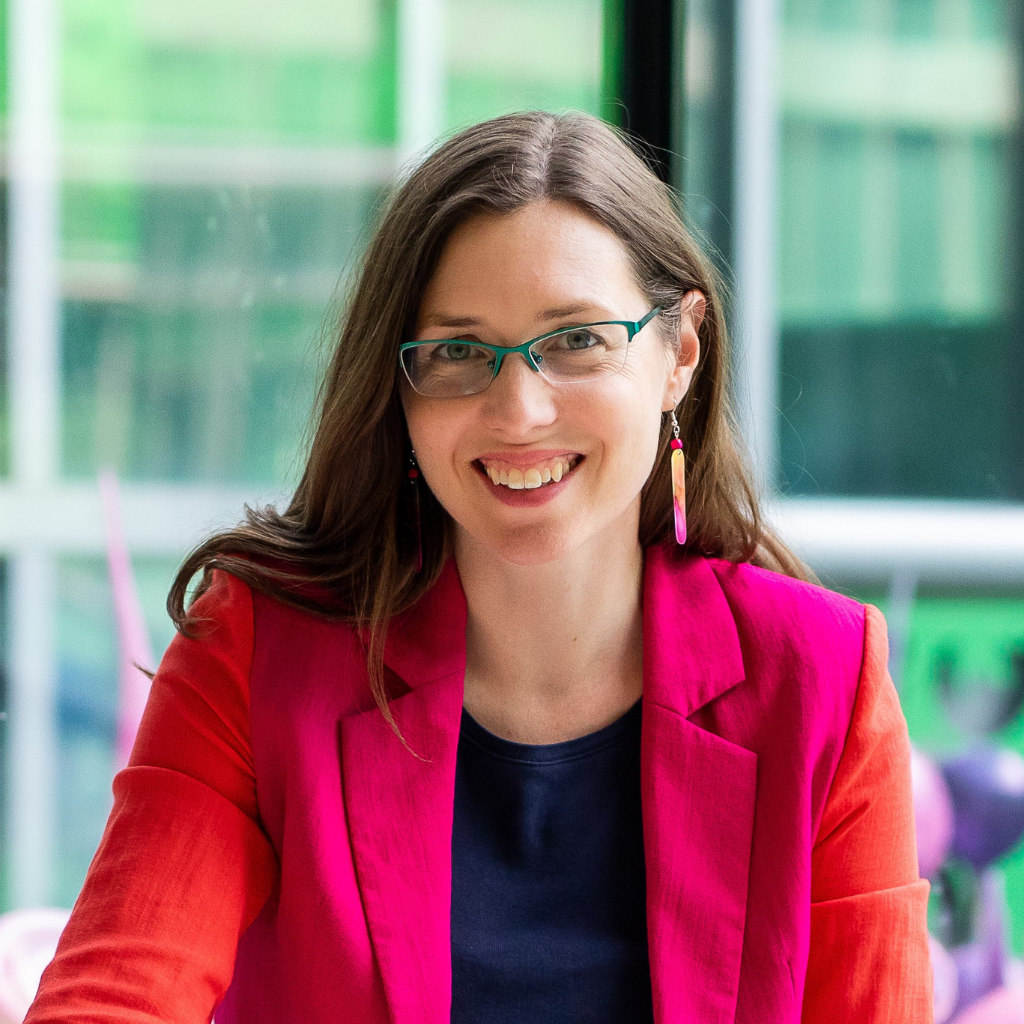 Photograph of Aubrey Reeves, a white woman with shoulder length brown hair. She is wearing green glasses and a pink suit.