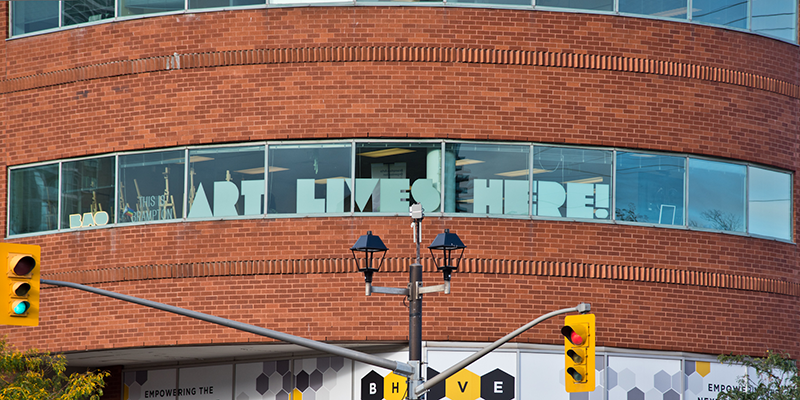 A brick building that is curved with a row of glass window in between the top and bottom rows of bricks with a sign in the windows saying "ART LIVES HERE"