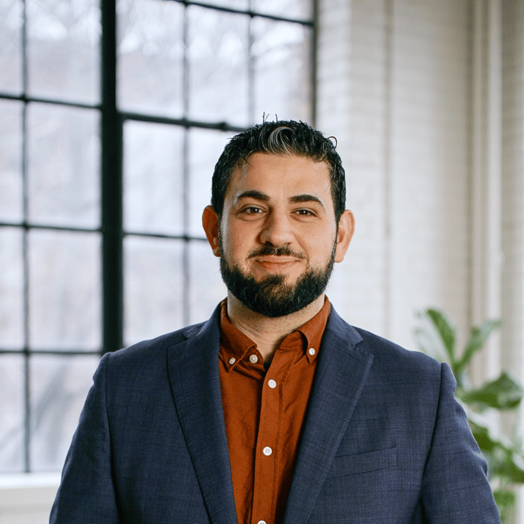 Photograph of a white man with a neatly trimmed beard. He is wearing a blue blazer and a brown button down shirt.