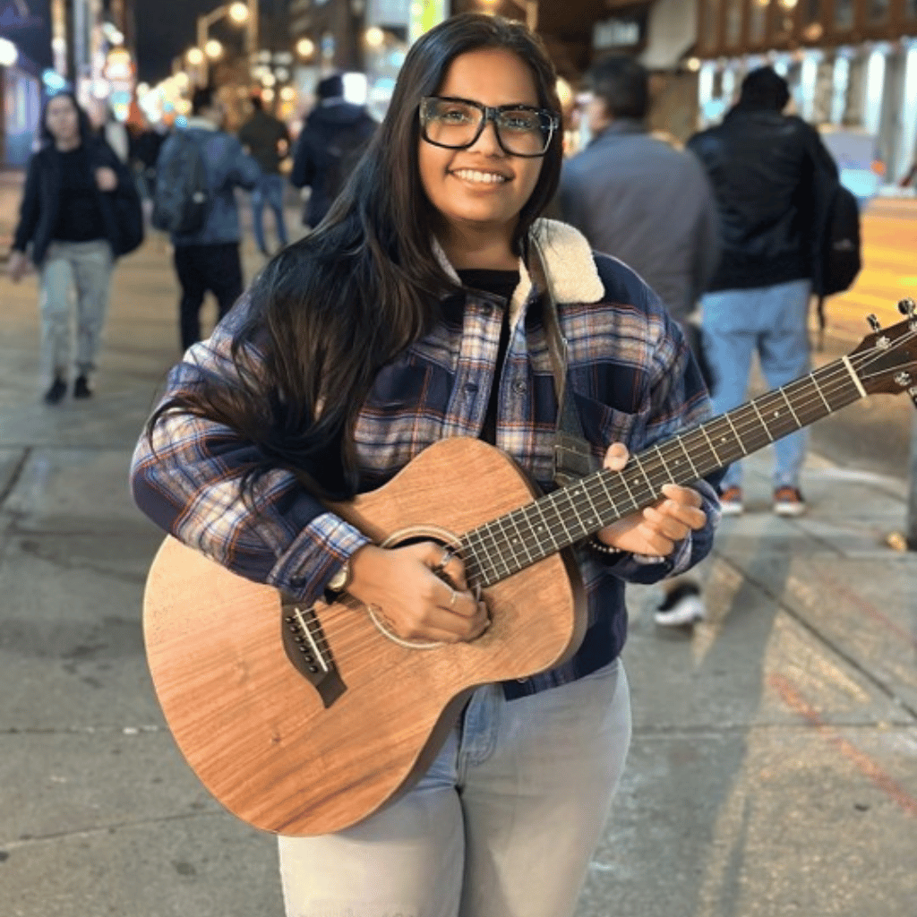 Photograph of a South Asian woman standing on a sidewalk while playing her guitar. She has long straight hair and is wearing black rimmed glasses, jeans and a blue plaid jacket.