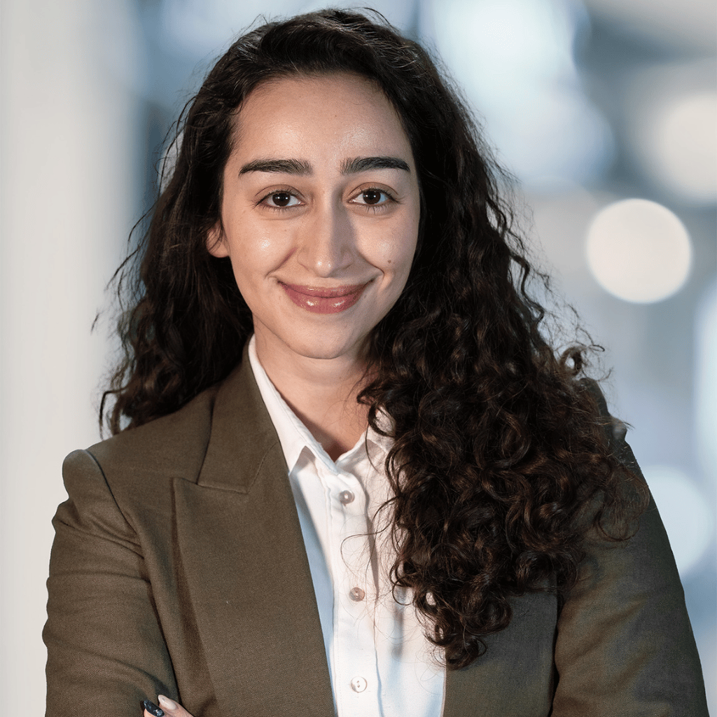 Photograph of a young woman of colour with long curly hair. She is wearing a brown blazer and a white button up shirt.