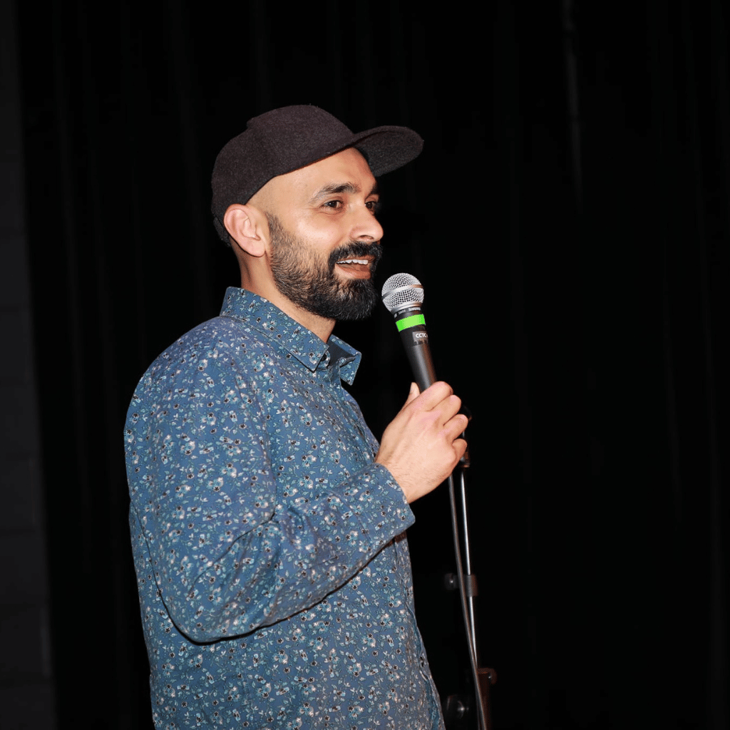 Photograph of a South Asian man with a neatly trimmed beard who is holding a mic up. He is wearing a black cap and a blue button up shirt shirt with a flower pattern on it.