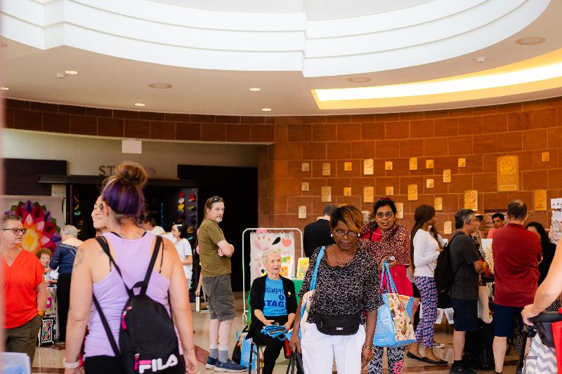 A crowd of people in summer outfits shopping around an art market inside a brick building with a white ceiling