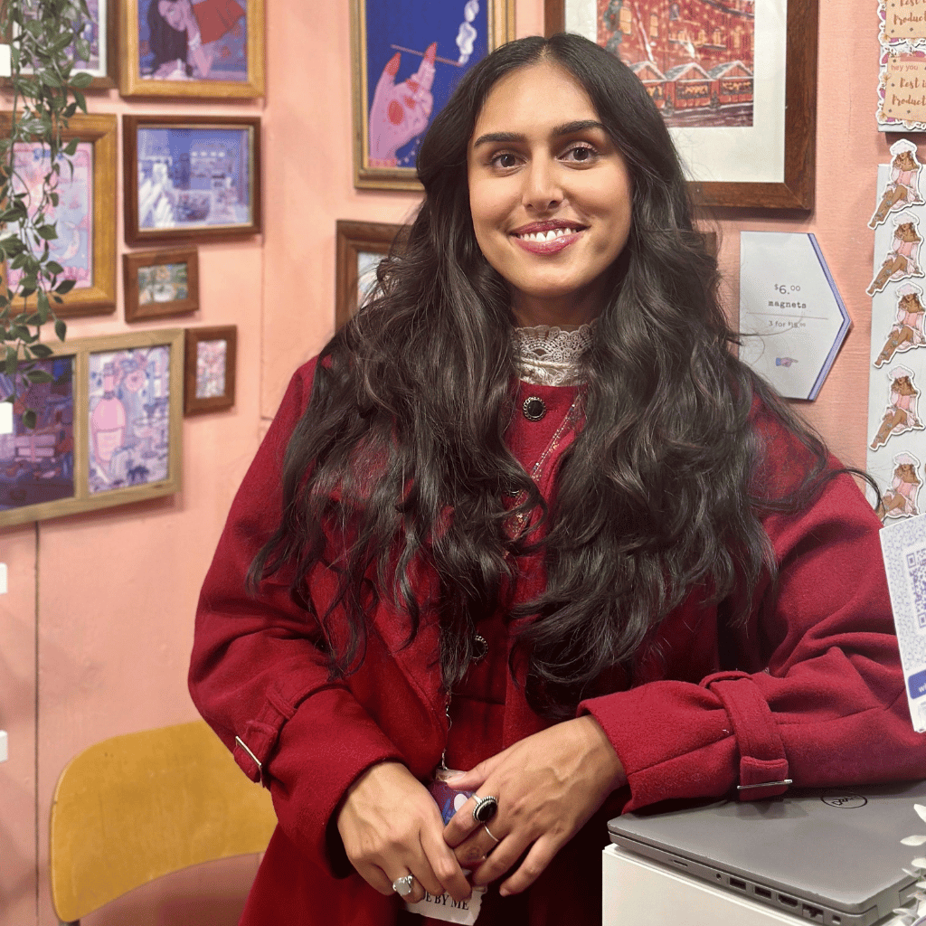 Photograph of Sabbie Narwal, a South Asian woman with long brown hair as she poses in her artist booth. She is wearing a auburn coat.