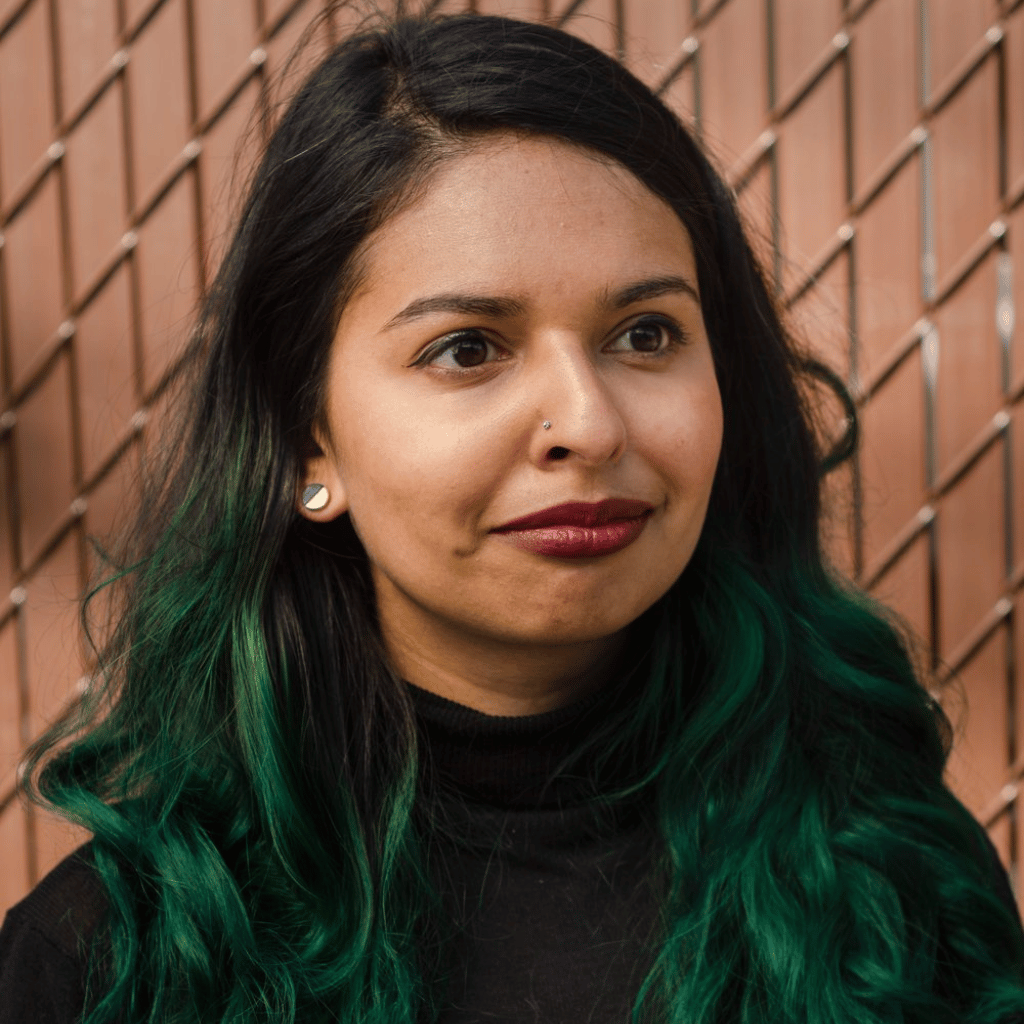 Photograph of a woman of colour with long wany hair. She is wearing a black turtleneck.