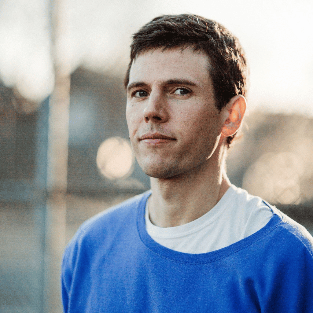 Photograph of Matthew Bailey, a white man with short brown hair, who is wearing a white t-shirt with a blue t-shirt over it.