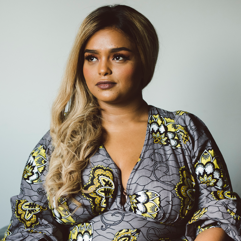 Photograph of Rania El Mugammar with long dyed blond hair and wearing a gray dress with yellow flower patterns on it.