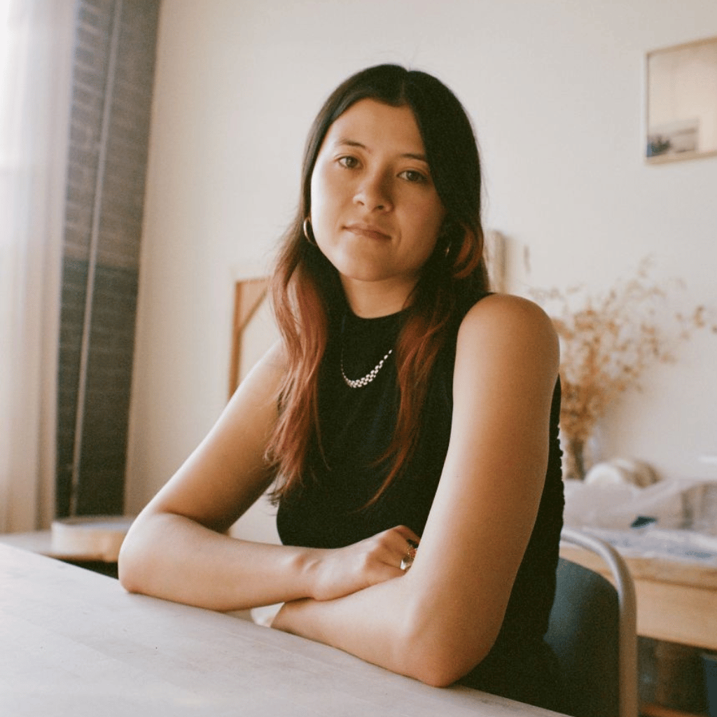Photo of Holly Chang, an East Asian woman with long brown hair. She is sitting at a table with her arms crossed. She is wearing a black dress and a silver necklace.