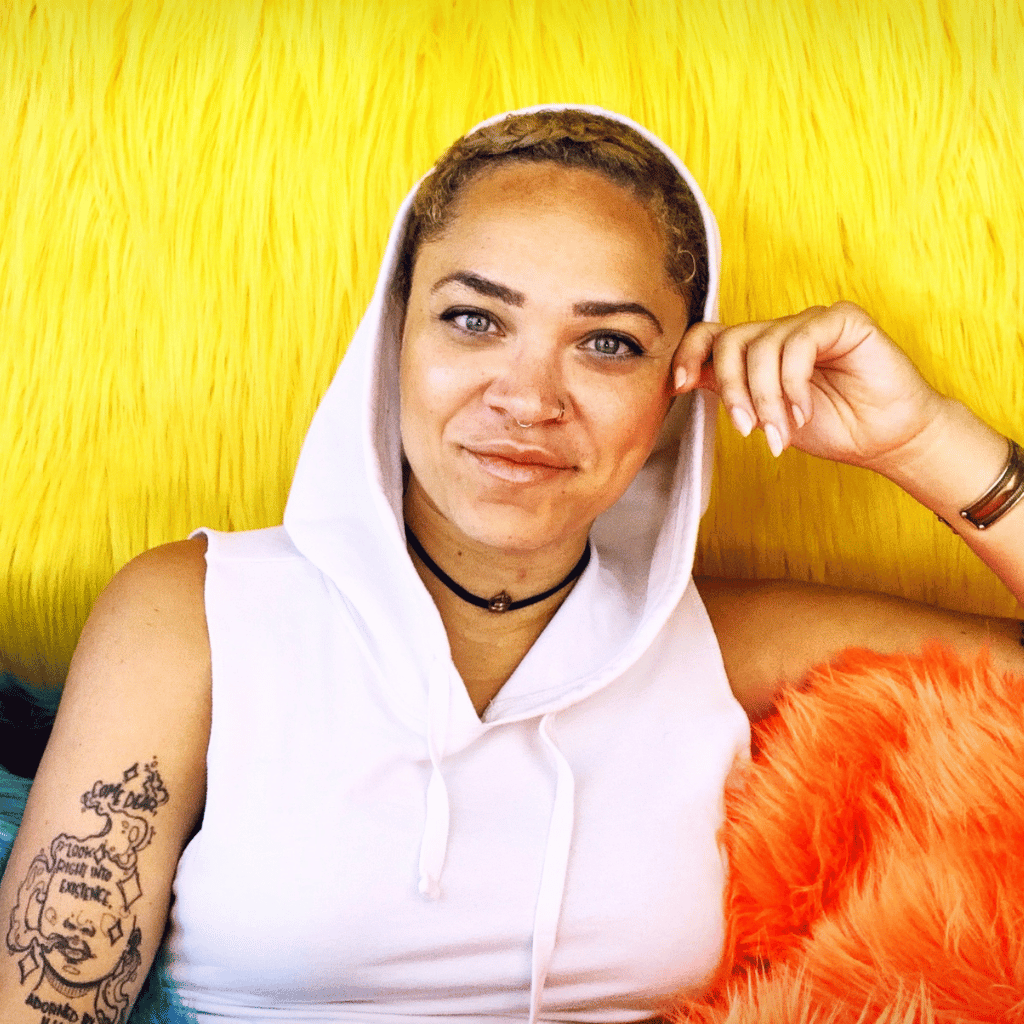 Photograph of Tasha Schumann, a Black woman with close-cropped hair. She is wearing a sleeveless white hoodie as she sits on an orange fur couch.