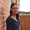 Photograph of Ankita Alemona, a South Asian woman in a navy blue patterned blouse leaning against a brick wall outdoors with sunlight in the background.
