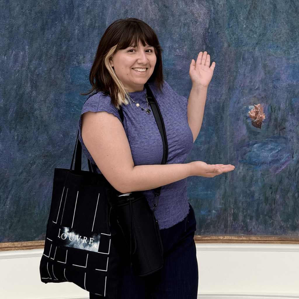 Photograph of Jasmine Canaviri, a Latinx woman with straight shoulder-length hair who is posing in front of a large painting. She is wearing a blue t-shirt and black pants.