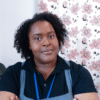 Photograph of Shiemara Hogarth, a Black woman with short curly hair and is wearing a black blouse with a blue apron over it.