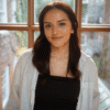 Photograph of the Canon Canada + Brampton Arts Organization Photographer in Residence Laya Mendizabal. She is a young woman with long brown hair wearing a black top and white striped shirt standing indoors near a window with natural light.
