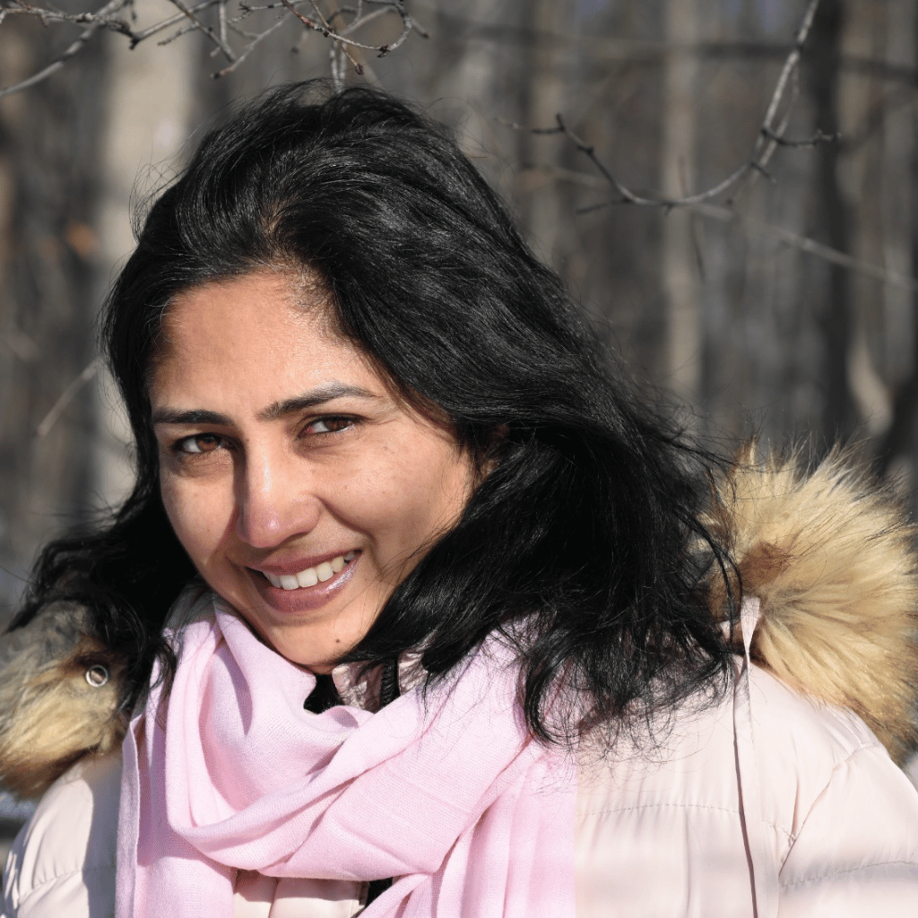 A South Asian woman wearing a light pink winter coat with a fur-trimmed hood and a matching pink scarf outdoors in a leafless forest.