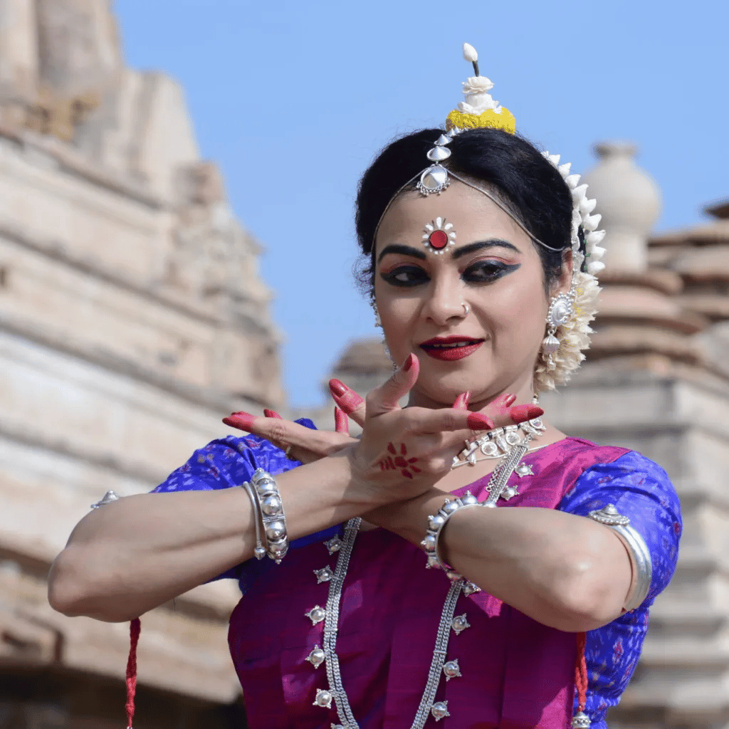 A South Asian woman in classical Indian dancer attire performing a hand gesture with temple architecture in the background.