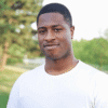 Photograph of Malachi Watson‑Narcisse, a Black wearing a plain white t-shirt standing outdoors with green trees blurred in the background.