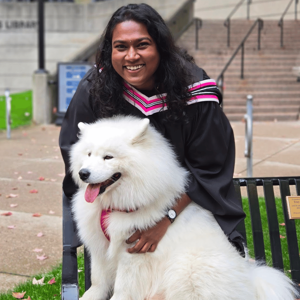 A Tamil woman in a black graduation gown with pink-striped collar sitting on a bench, hugging a large fluffy white Samoyed dog outdoors near steps.