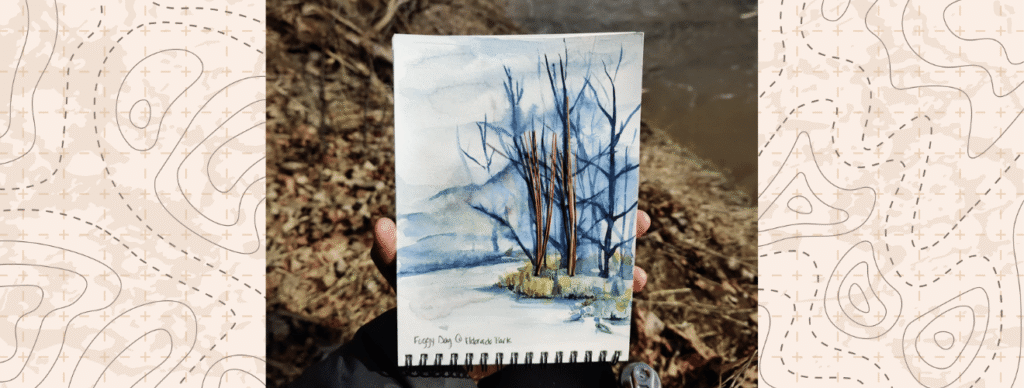 Photograph of a watercolour painting of trees on a journal being held by the artist in a ravine